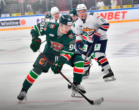 Хоккейный матч: игрок в зелёной форме с клюшкой ведёт шайбу, его преследуют двое соперников в белой форме. Яркая динамика на льду, действие разворачивается возле баннеров с рекламой